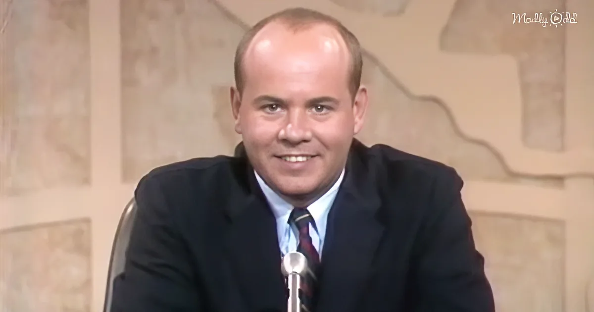 A man in a suit and tie smiles while sitting in front of a microphone, with a decorative wall behind him.