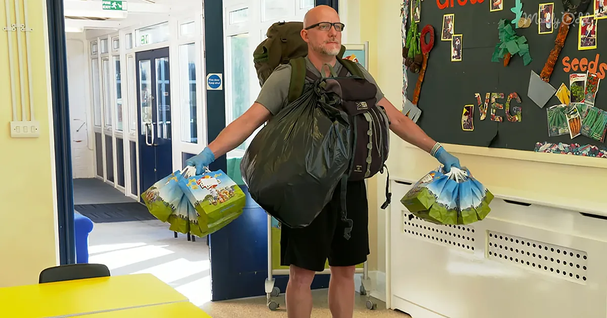 A man with a backpack and gloves holds bags of supplies in a classroom.
