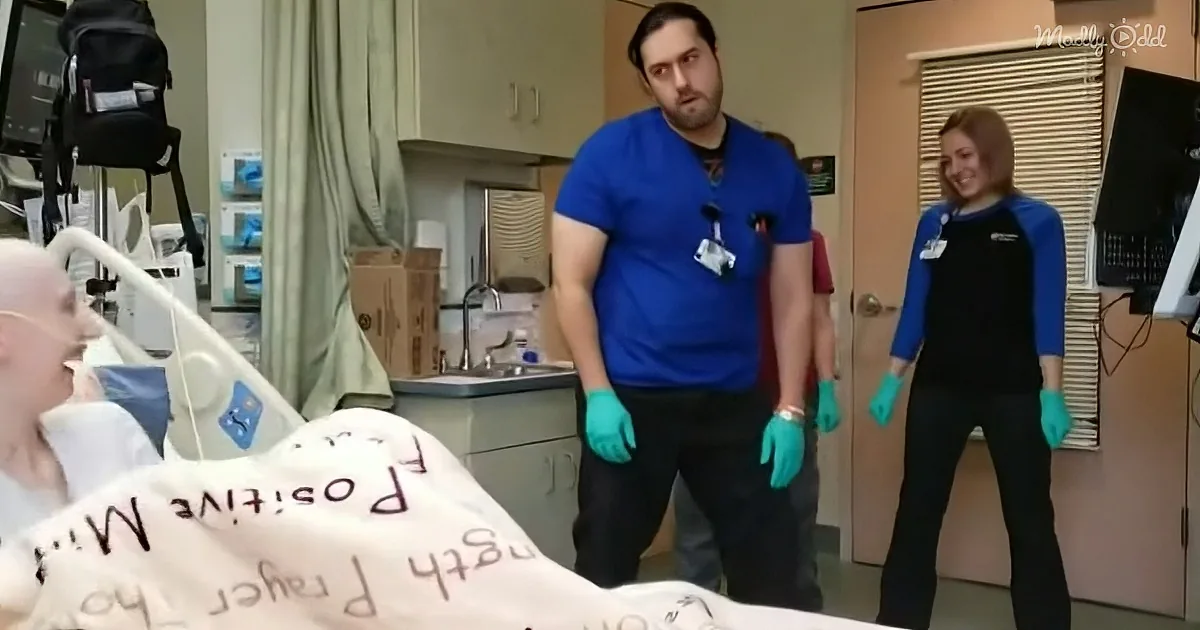 A man in blue scrubs and gloves stands beside a hospital bed, interacting with a patient.
