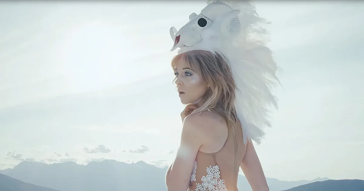 A woman in a white dress with a lion headdress stands against a mountainous backdrop.