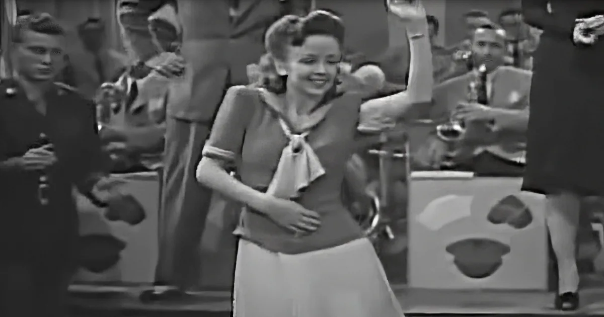 A woman in a sailor-style dress raises her arm, smiling, with a band and audience in the background.