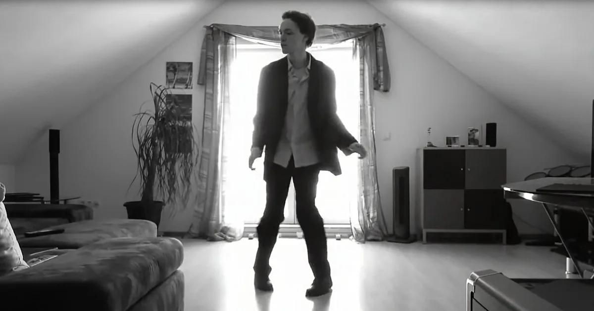 A person stands in a room with a slanted ceiling, wearing a jacket and pants, near a window with curtains.