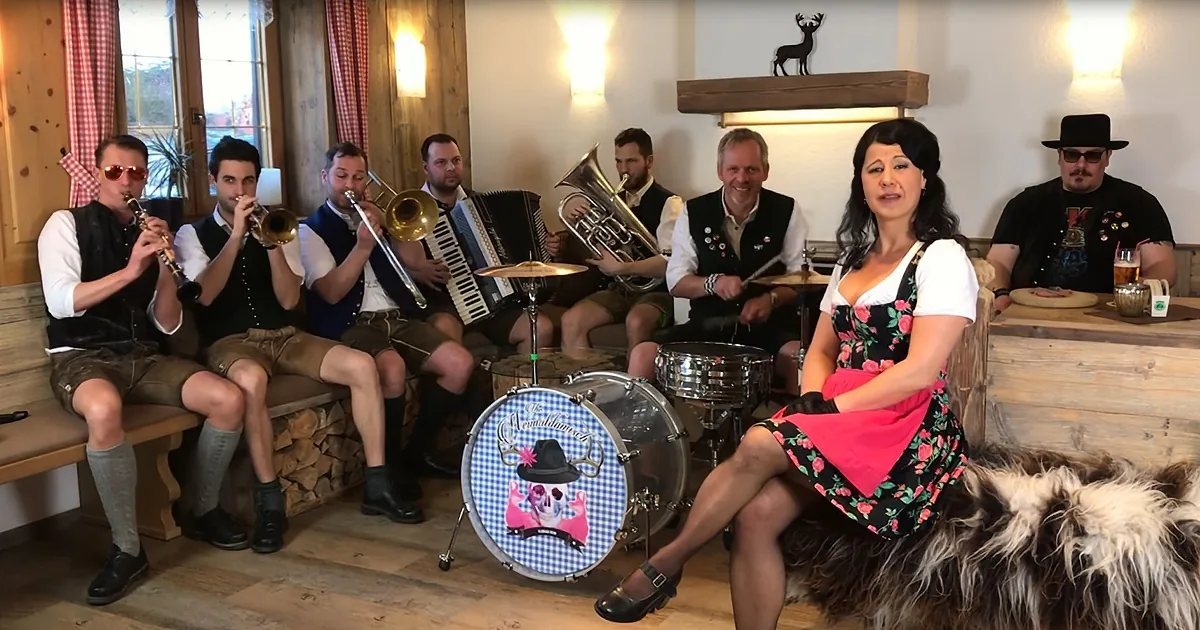 A woman in a dirndl sits on a bench, smiling, with a band playing traditional instruments behind her.