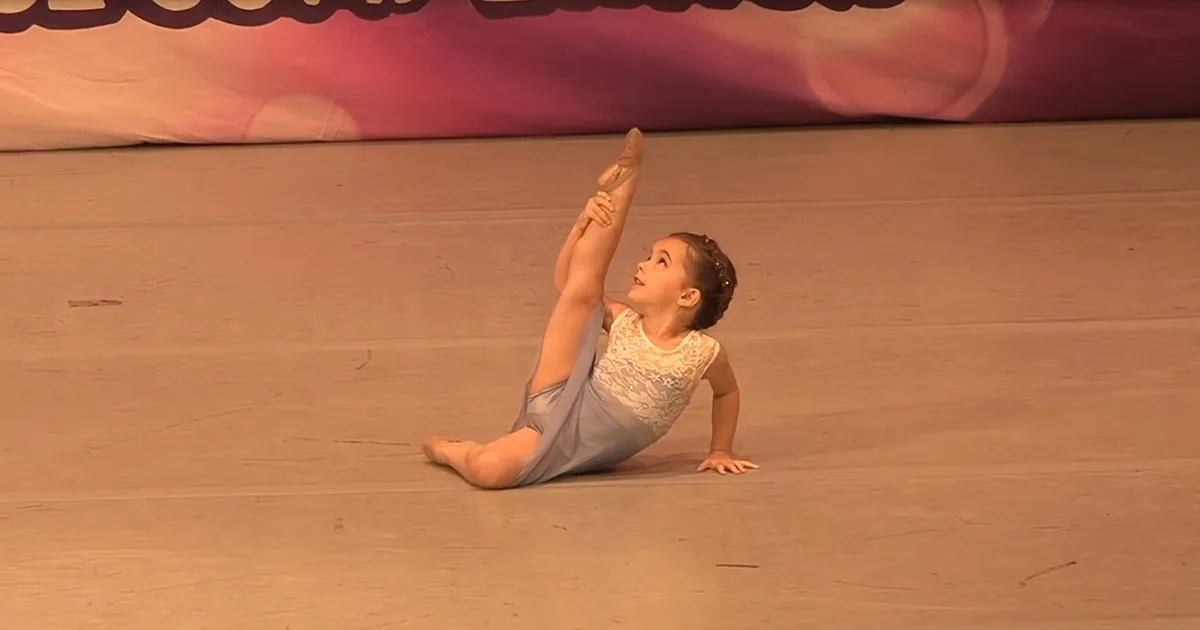 A young girl in a ballet pose, holding her leg up while sitting on the floor.