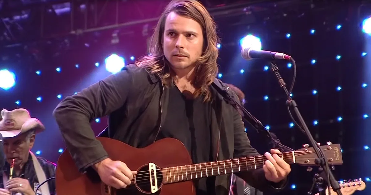 A man with long hair plays an acoustic guitar on stage, illuminated by blue lights.