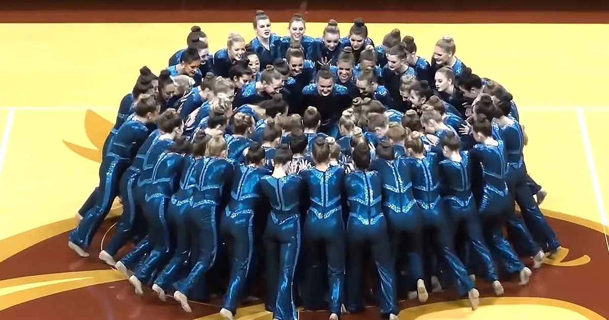 A group of synchronized gymnasts in blue outfits form a tight huddle on a yellow and brown court.