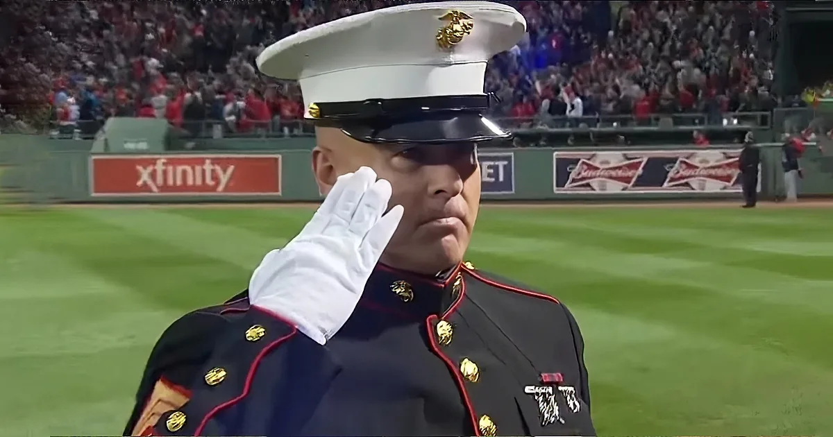 A Marine in uniform salutes on a baseball field with a crowd and advertisements in the background.