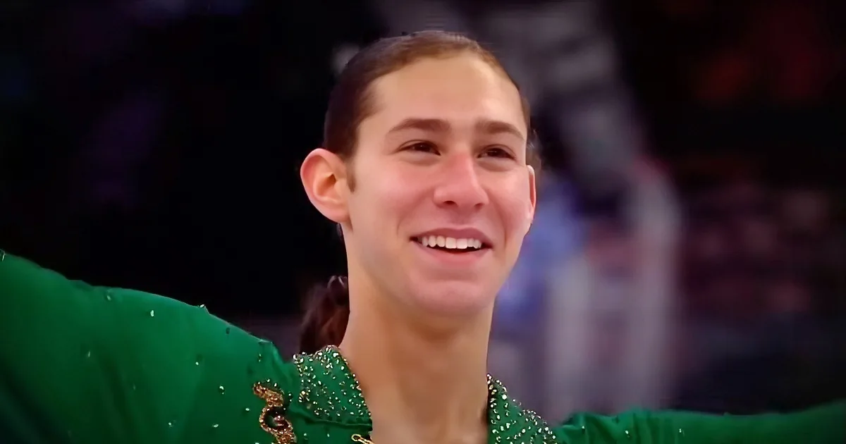 A person in a green outfit with sparkling details smiles, arms outstretched, against a blurred background.