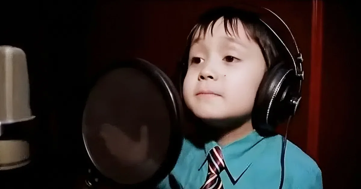 A child wearing headphones and a tie stands in front of a microphone, likely recording or singing.