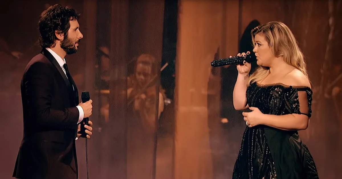 A man in a suit and a woman in a black dress sing into microphones on stage.