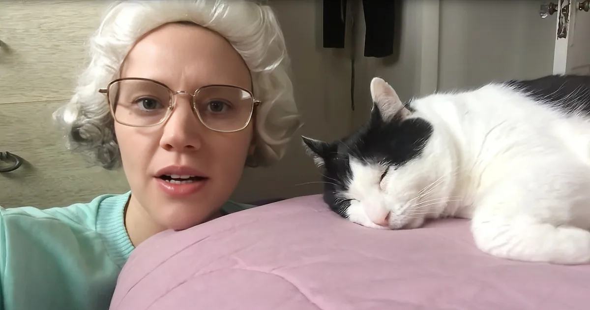 A person in a wig and glasses sits beside a sleeping black-and-white cat on a pink surface.
