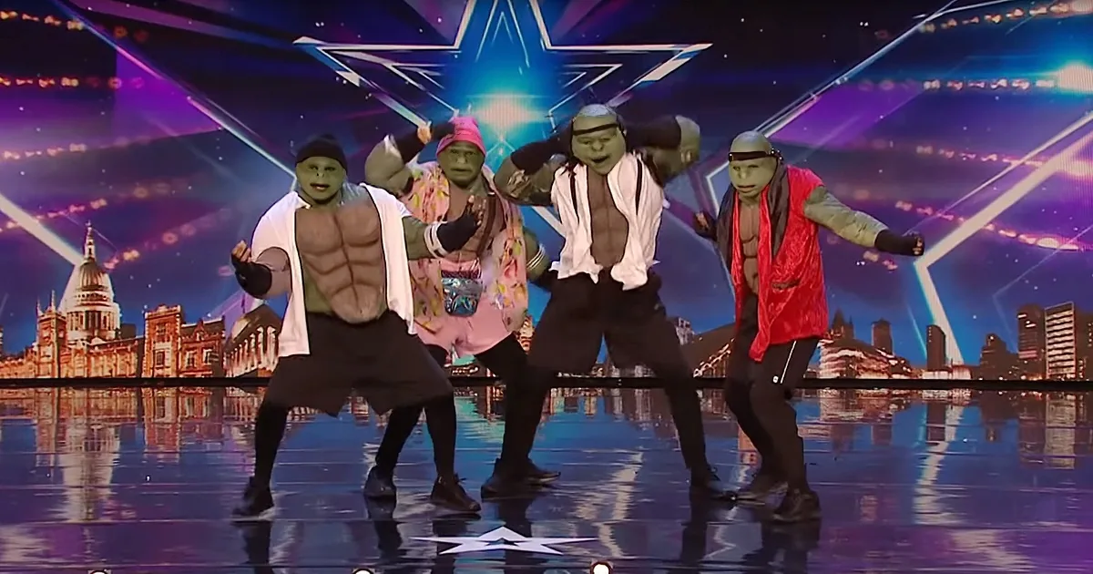Four performers in green alien costumes dance on a stage with a cityscape backdrop and a star-shaped light.
