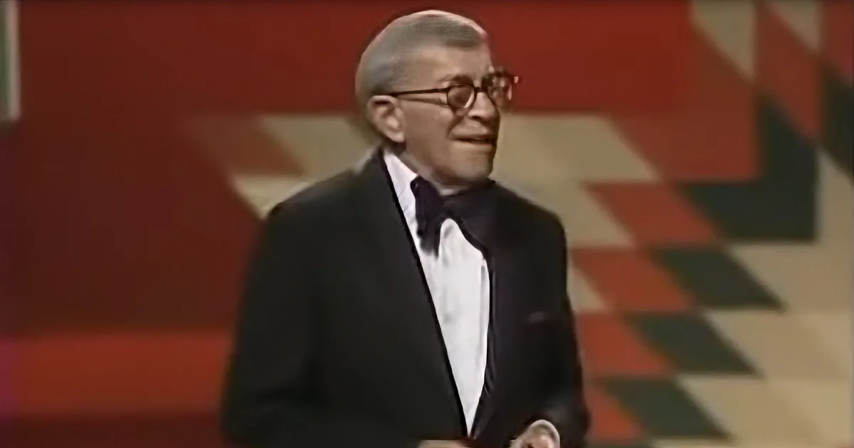 A man in a tuxedo and bow tie smiles while holding a microphone, standing in front of a red and beige patterned background.