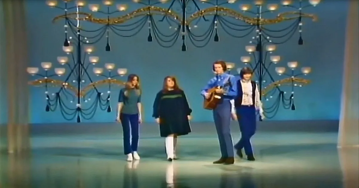 Four people in blue attire walk on stage under ornate chandeliers.
