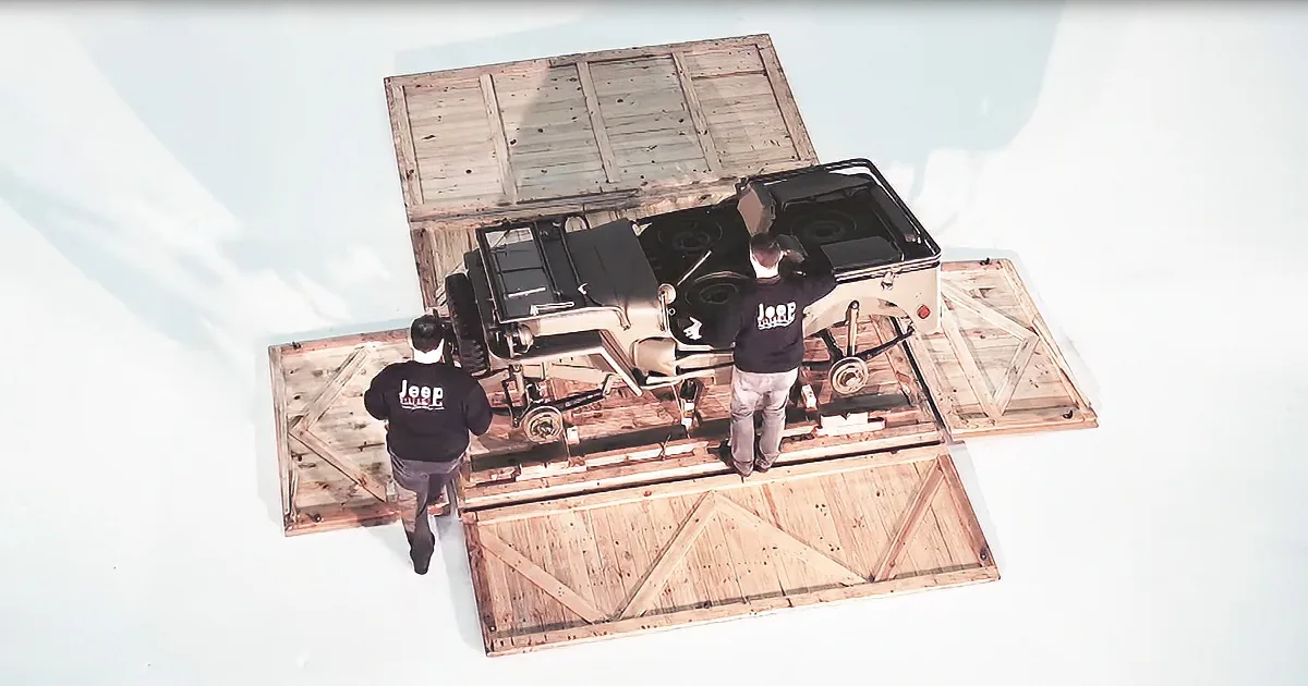 Two men in Jeep shirts work on a vintage Jeep on a wooden platform.