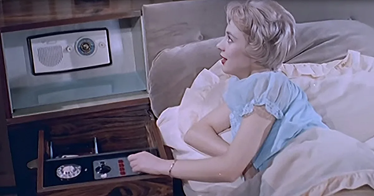 A woman in a blue nightgown lies in bed, interacting with a vintage radio and record player.