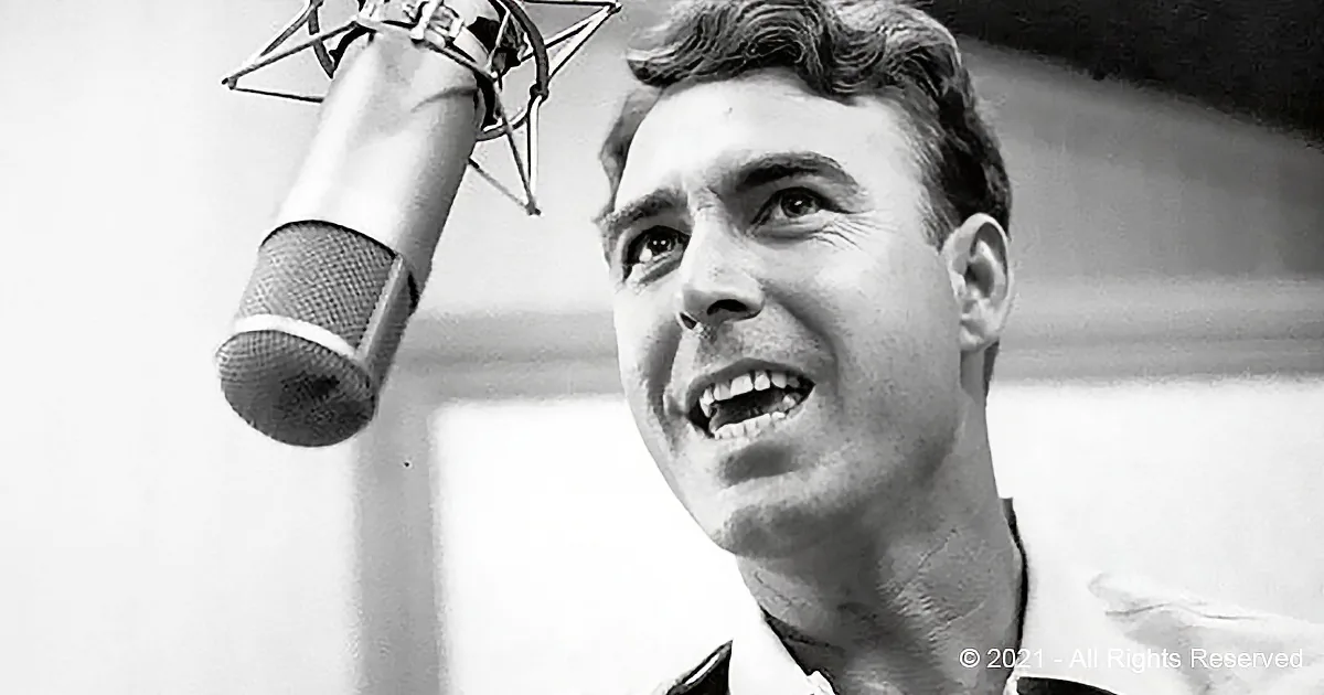 A man singing into a microphone, wearing a collared shirt, in a recording studio setting.