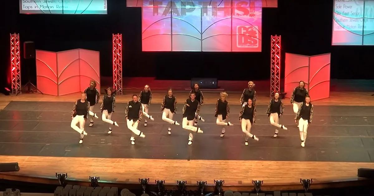 A group of dancers in black and white outfits perform a synchronized tap routine on a stage.