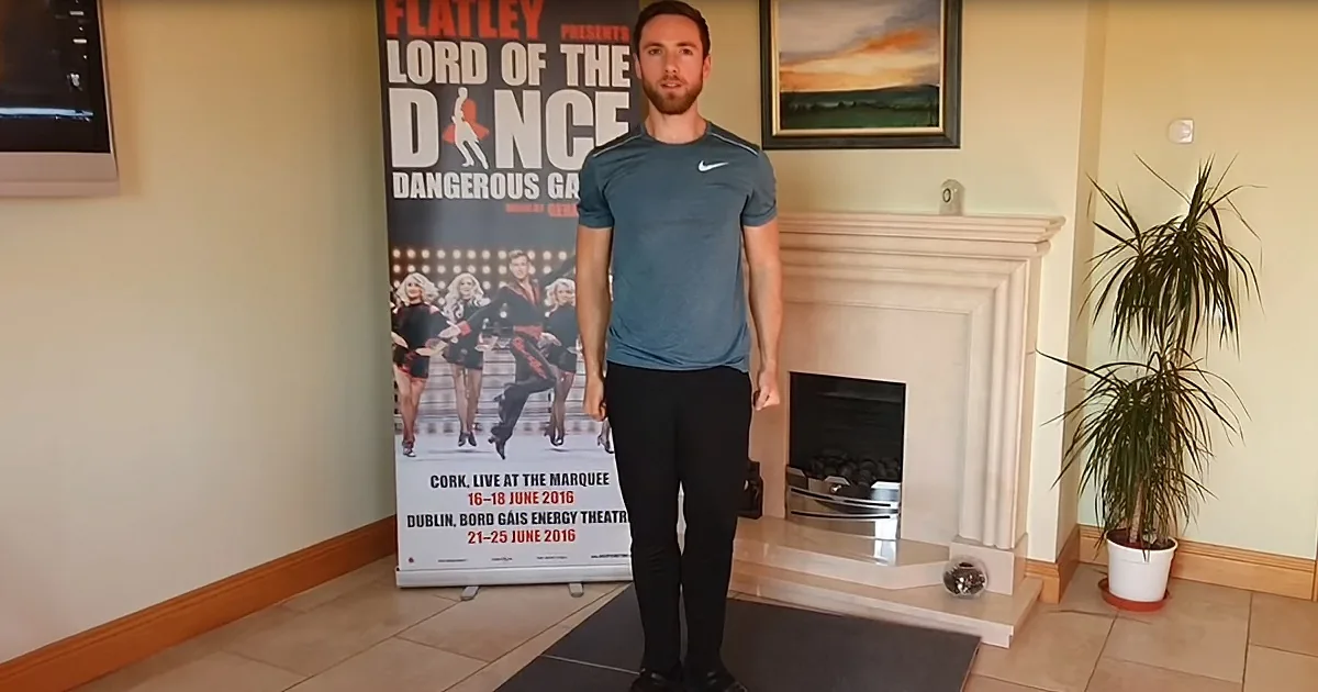 A man stands in front of a "Lord of the Dance" poster, wearing a Nike shirt and black pants, in a room with a fireplace...