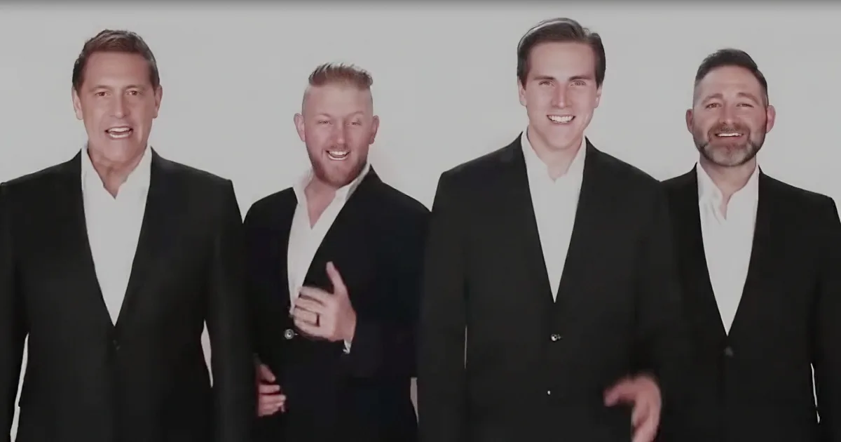 Four men in black suits and white shirts are smiling and posing together.