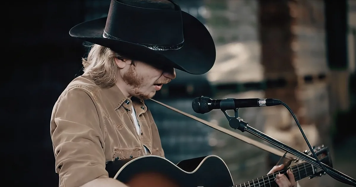 A man in a cowboy hat and tan shirt plays an acoustic guitar and sings into a microphone.