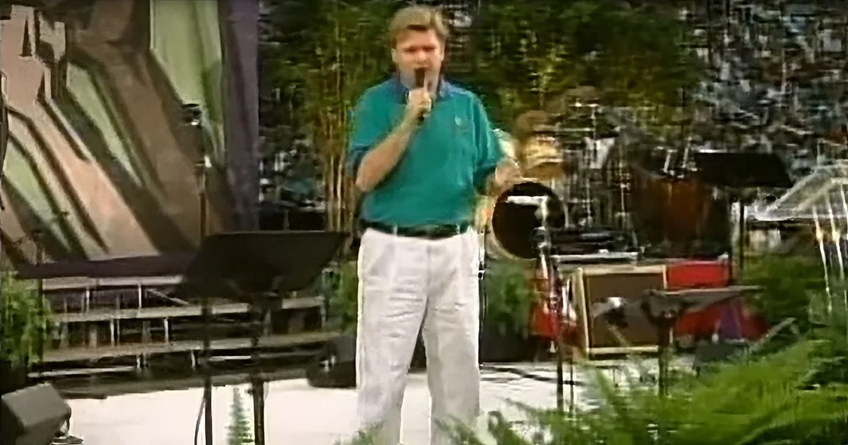 A man in a teal shirt and white pants stands on stage holding a microphone, surrounded by musical equipment and greenery.
