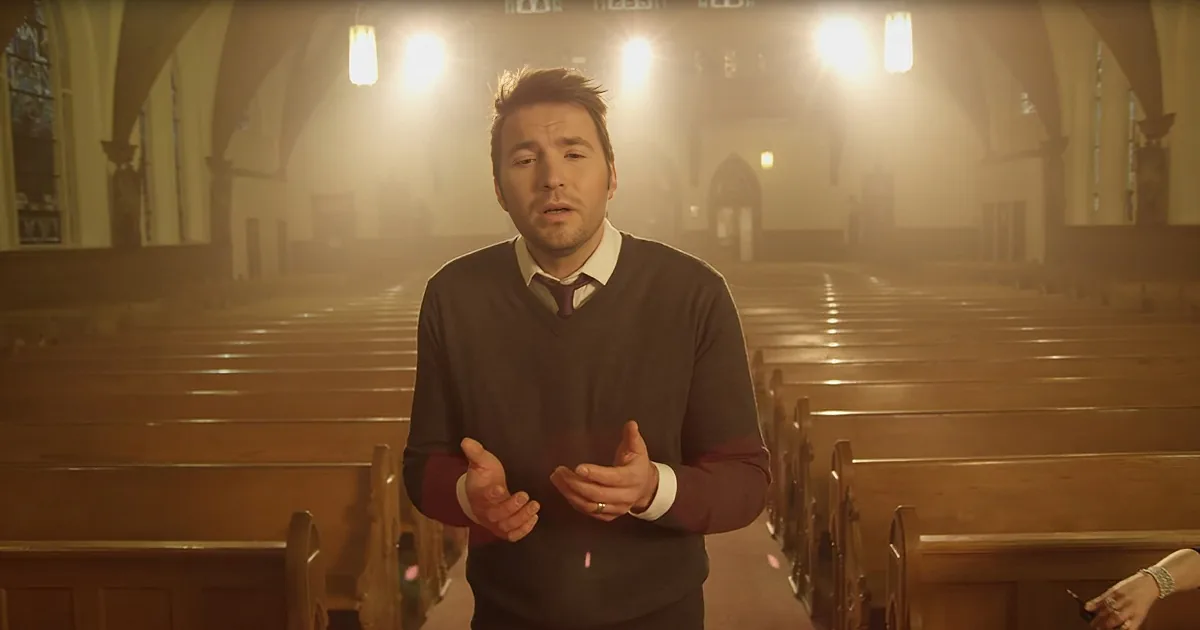 A man in a church, gesturing with hands, wearing a sweater and tie, surrounded by rows of pews.