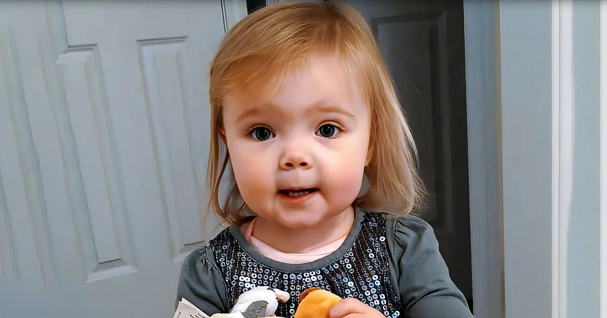 A young child with blonde hair and blue eyes, wearing a green sequined top, holds a stuffed animal and a piece of food.
