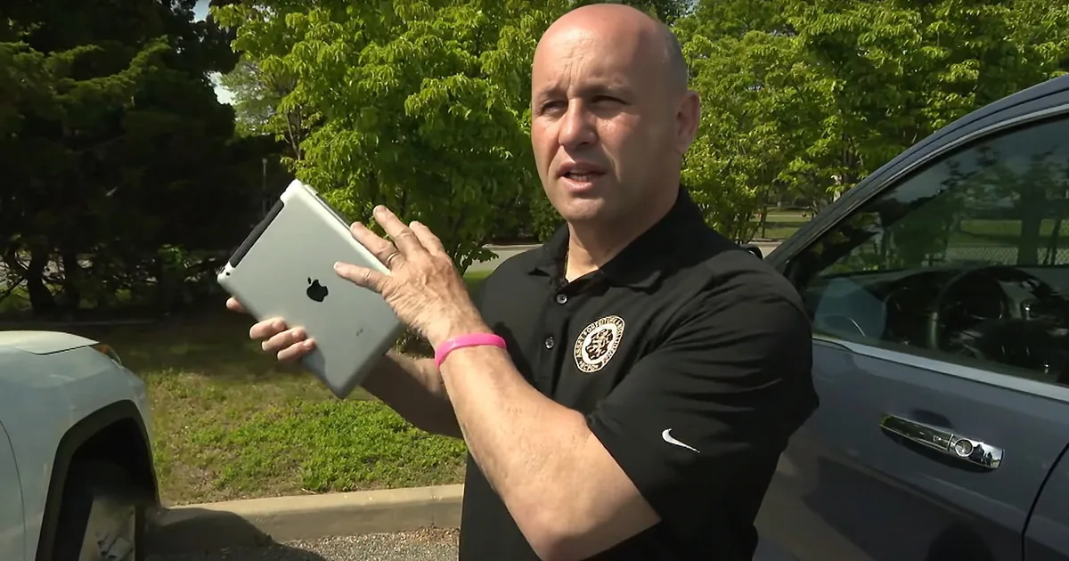 A man in a black polo shirt holds an iPad, gesturing with his other hand, standing outdoors near parked cars.