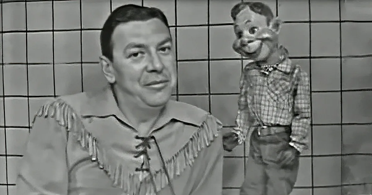 A man in a fringed shirt stands beside a puppet in a checkered shirt and jeans.