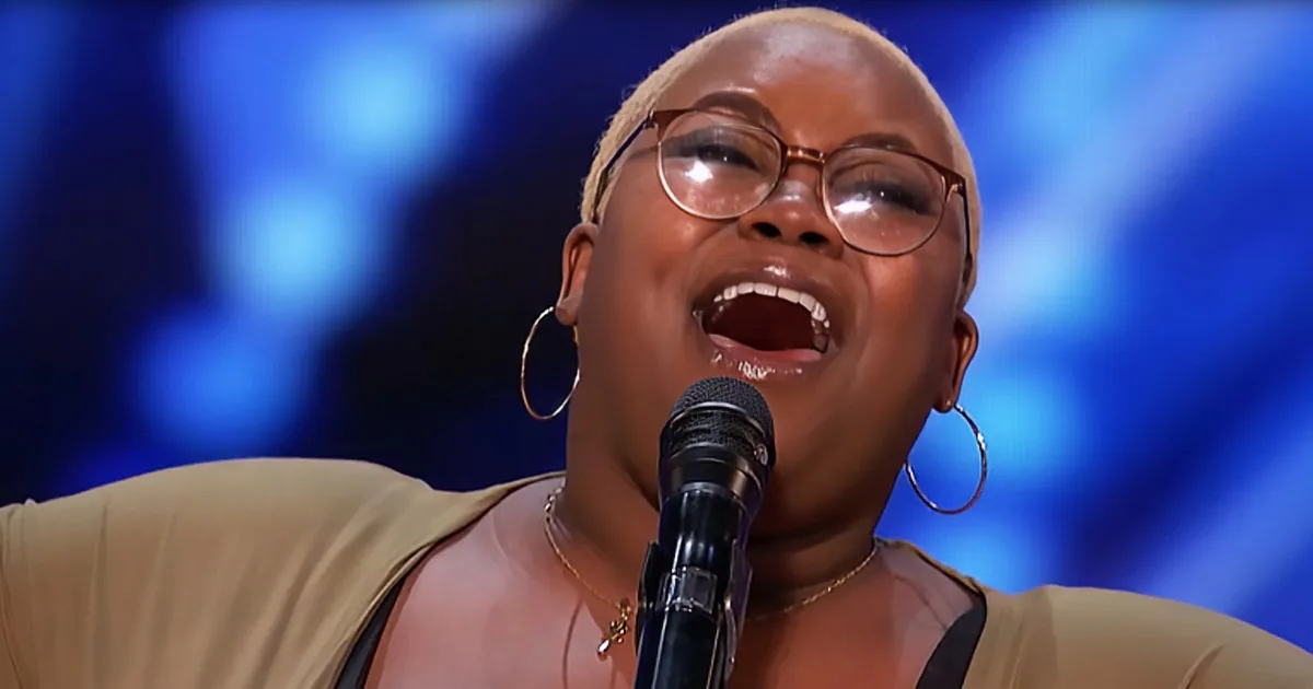A woman with glasses and hoop earrings sings into a microphone, wearing a beige top, against a blue background.