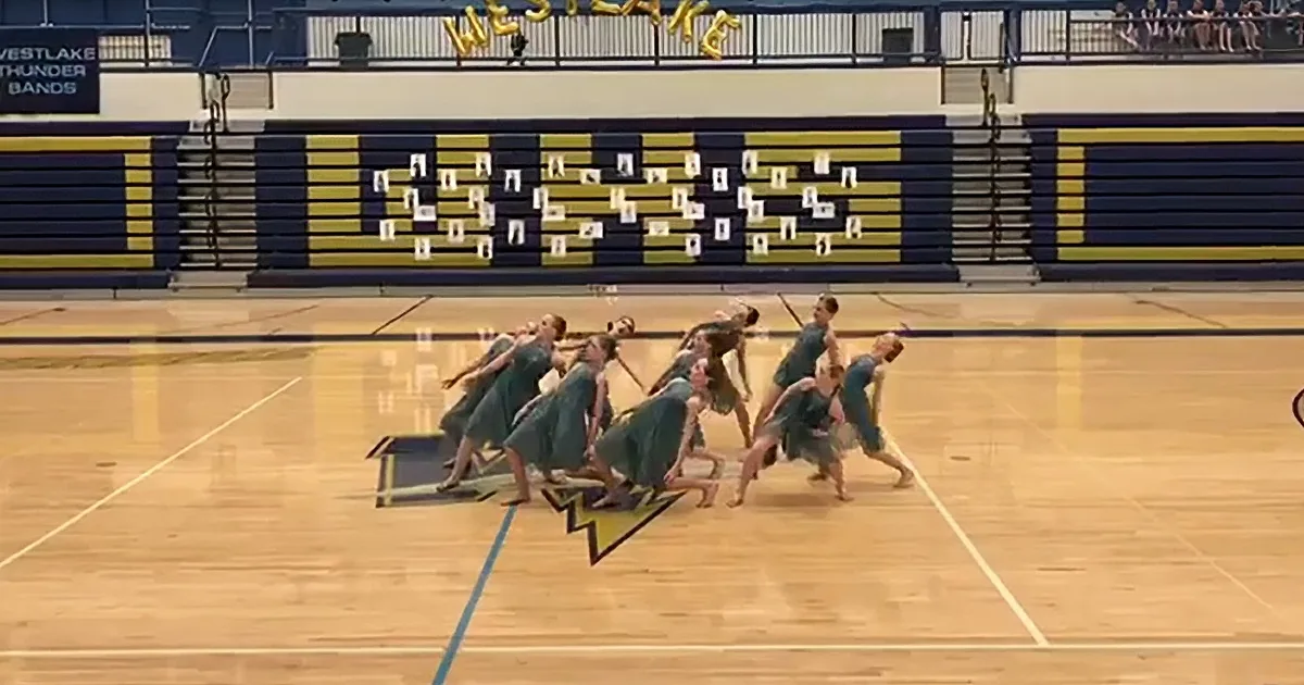 A group of dancers in teal dresses perform a synchronized routine on a gymnasium floor.