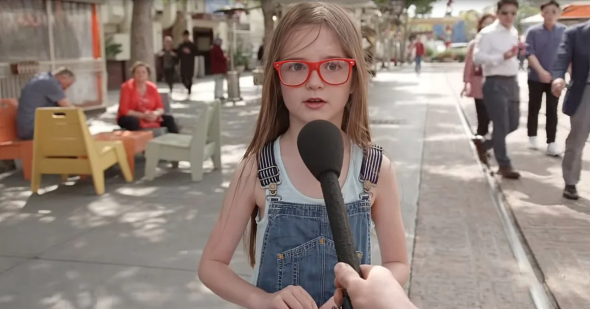 A young girl wearing red glasses and overalls is being interviewed on a sunny sidewalk.
