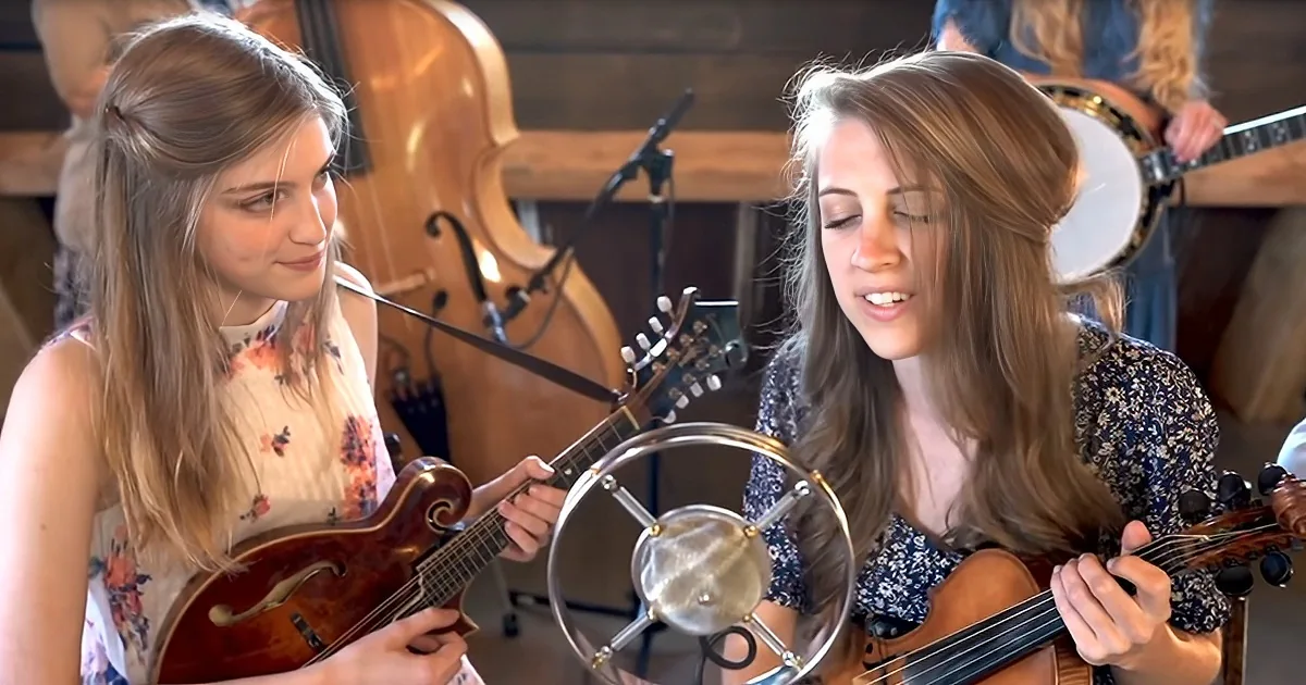 Two women play mandolin and violin in front of a microphone, with a cello and banjo in the background.