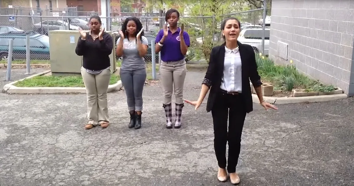 Four women stand outdoors, one in a blazer gesturing, others in casual attire, with a fenced area and parked cars in the...