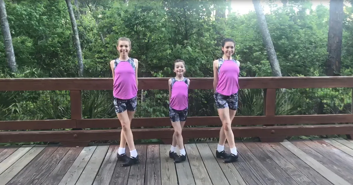Three girls in matching pink tops and black shorts stand on a wooden bridge, smiling.