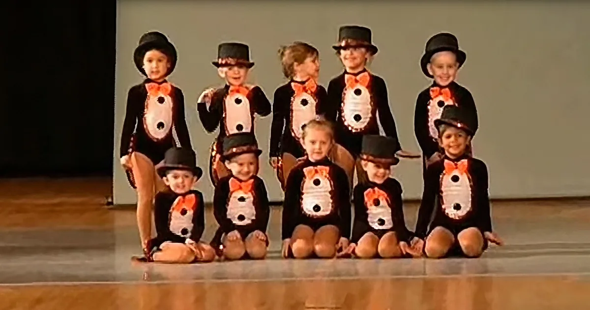 Children in penguin costumes with top hats pose on stage.