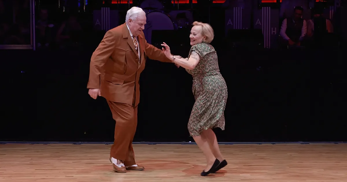 An elderly couple dances joyfully on a wooden floor, holding hands and smiling.