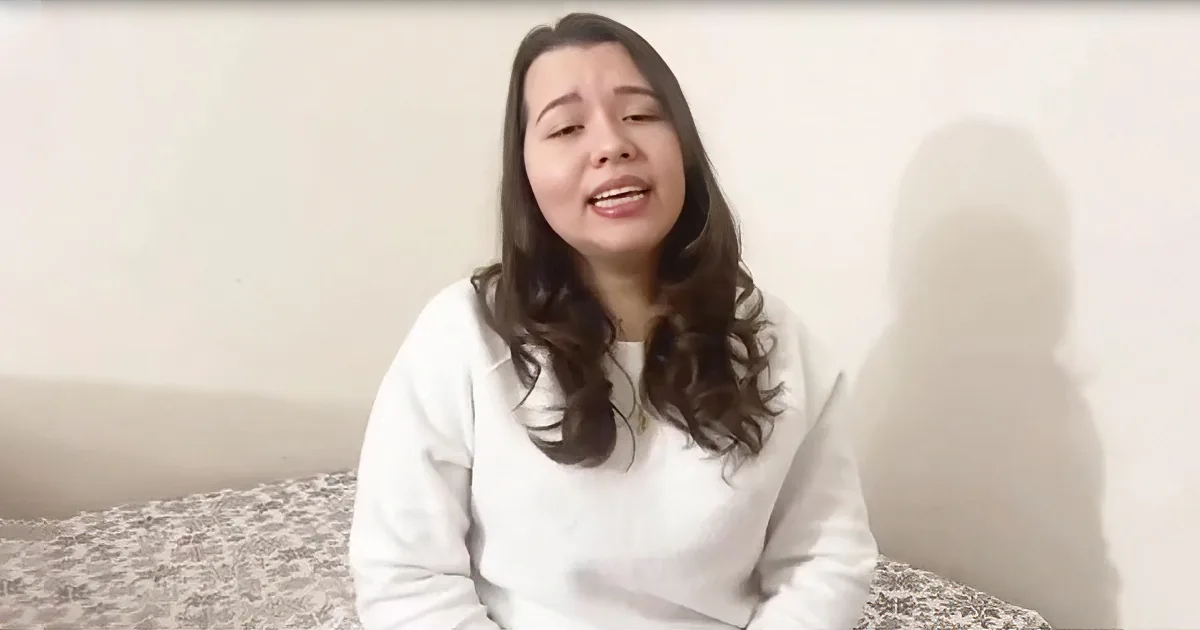 A woman with long dark hair, wearing a white top, sits on a patterned surface against a plain wall.