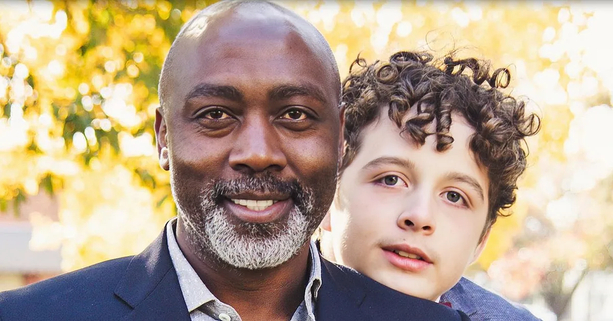 A man with a beard and a young boy with curly hair stand together, smiling, against a blurred autumnal background.