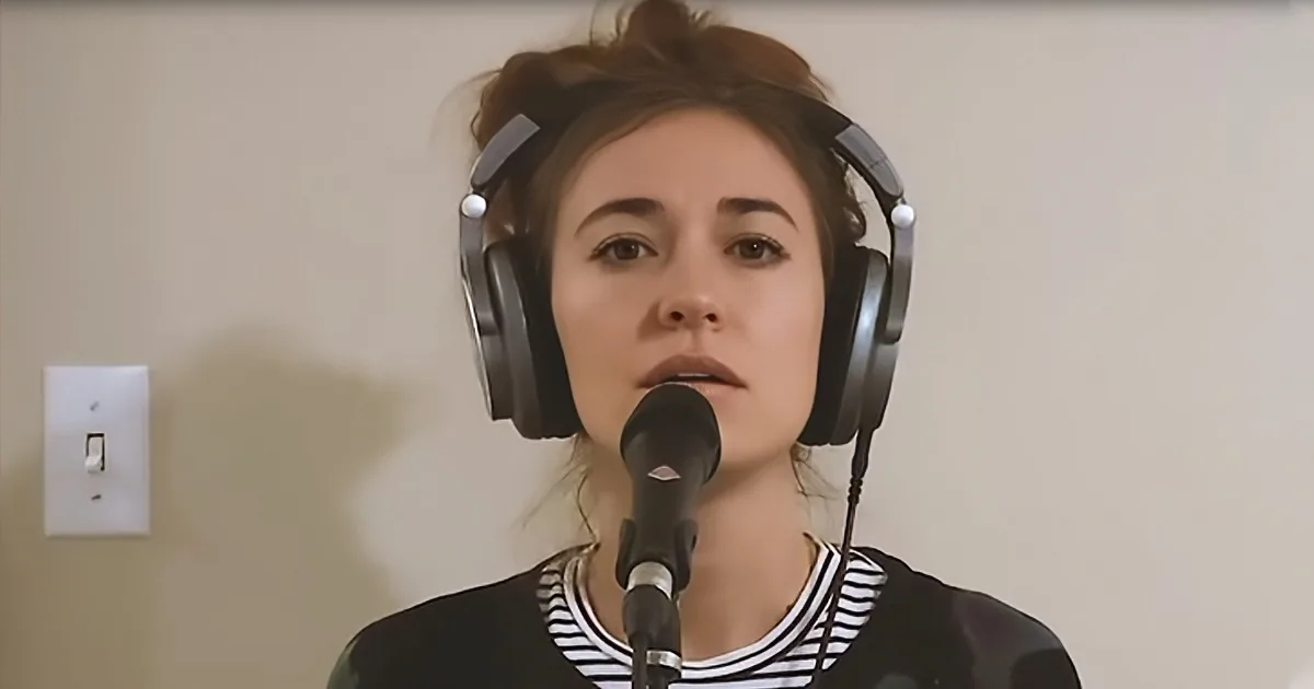 A person wearing headphones and a striped shirt speaks into a microphone in a room with a light switch on the wall.