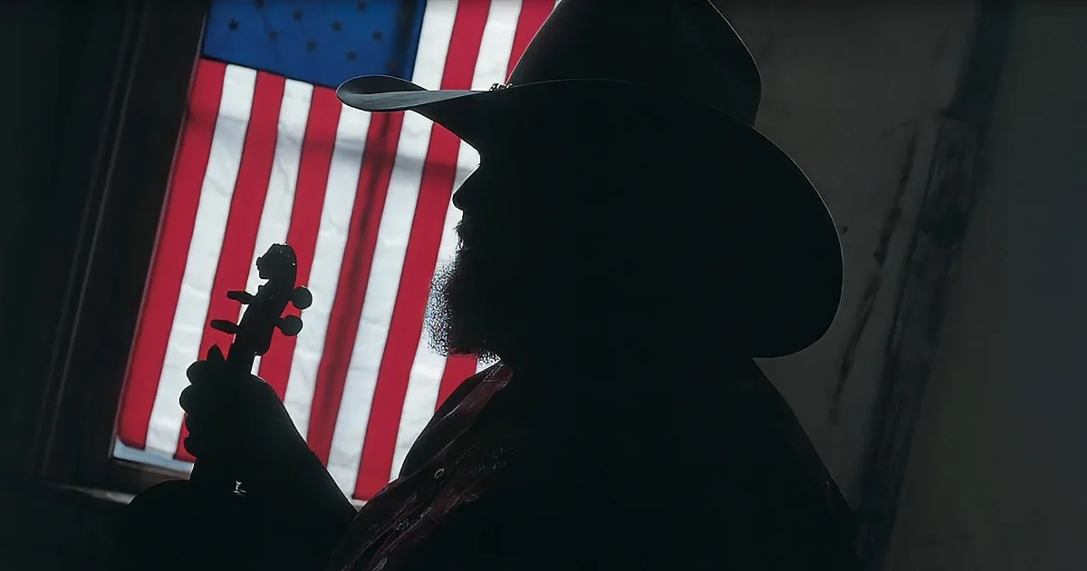 A silhouette of a person with a beard and hat holding a violin, with an American flag in the background.