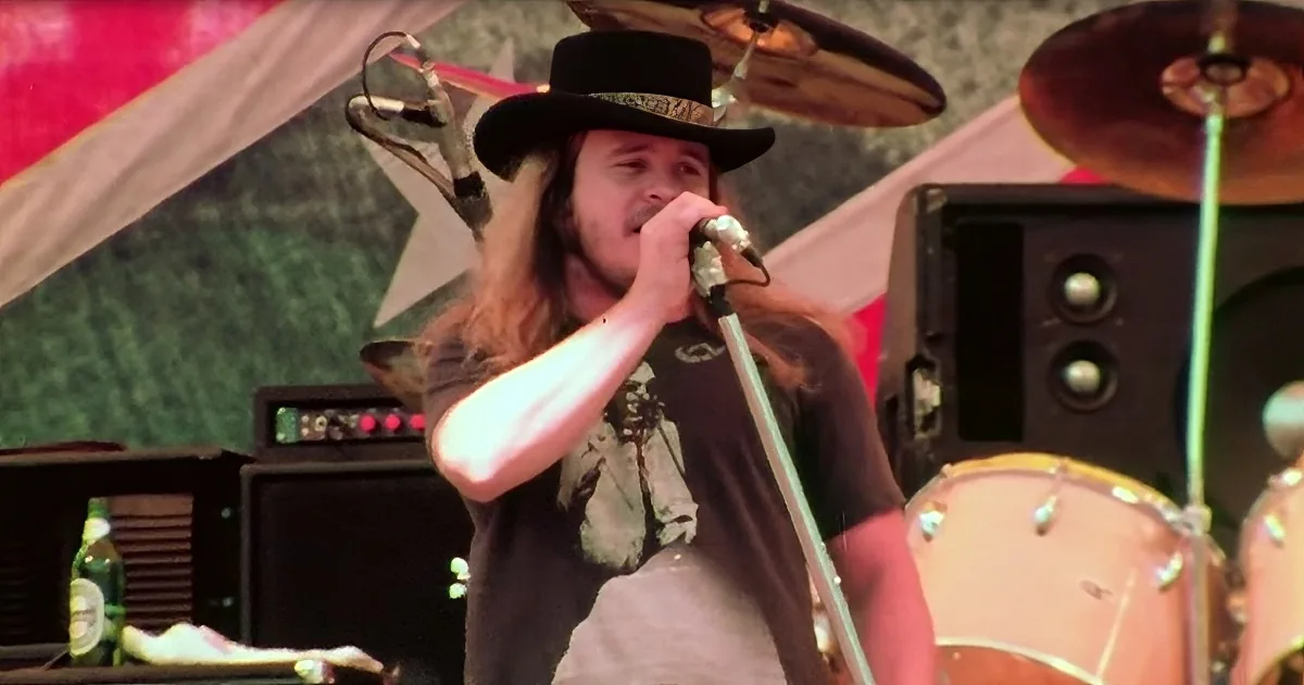 A person with long hair and a hat sings into a microphone on stage with a drum set and amplifiers behind them.