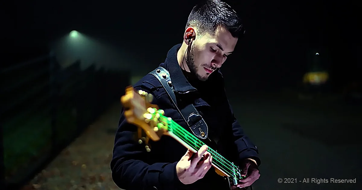 A man in a dark coat plays a bass guitar under dim lighting.