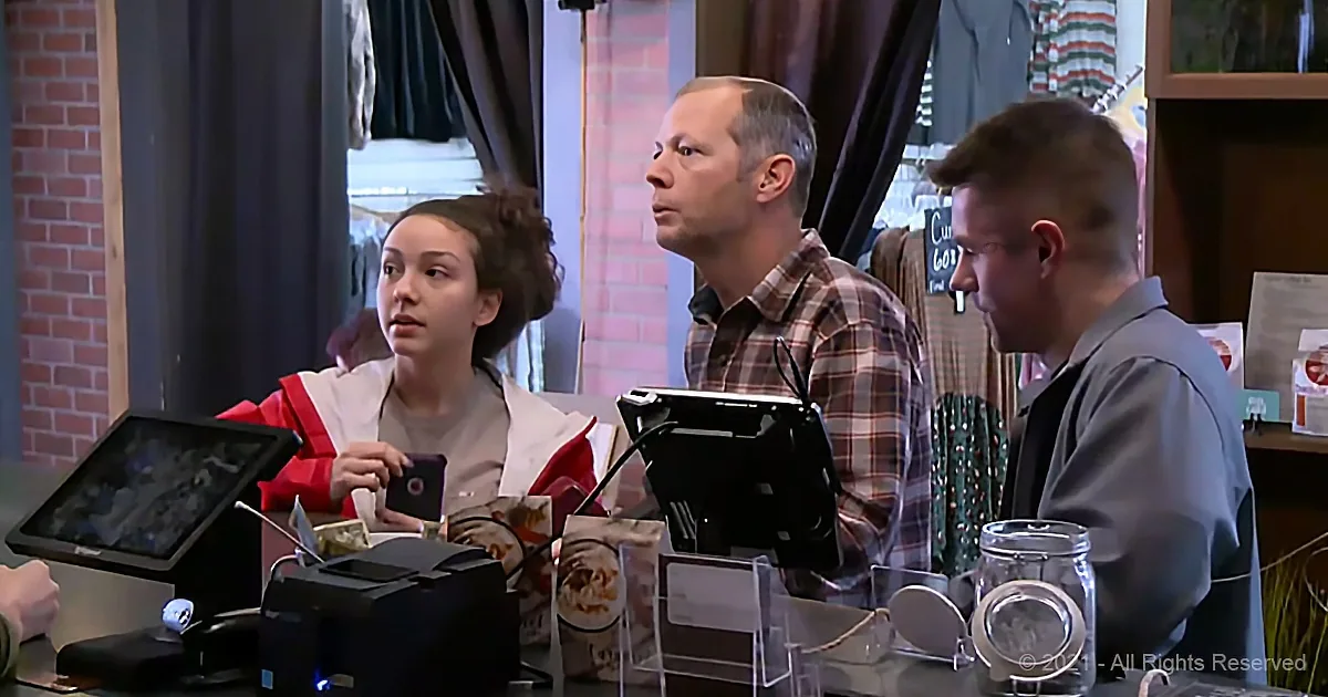 Three people at a checkout counter, one holding a phone, with a brick wall and clothing in the background.