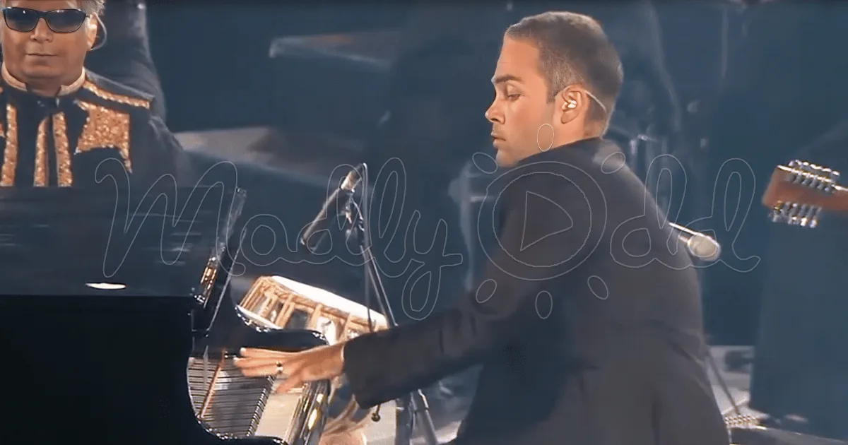 A man in a suit plays a piano, with a person in sunglasses in the background.