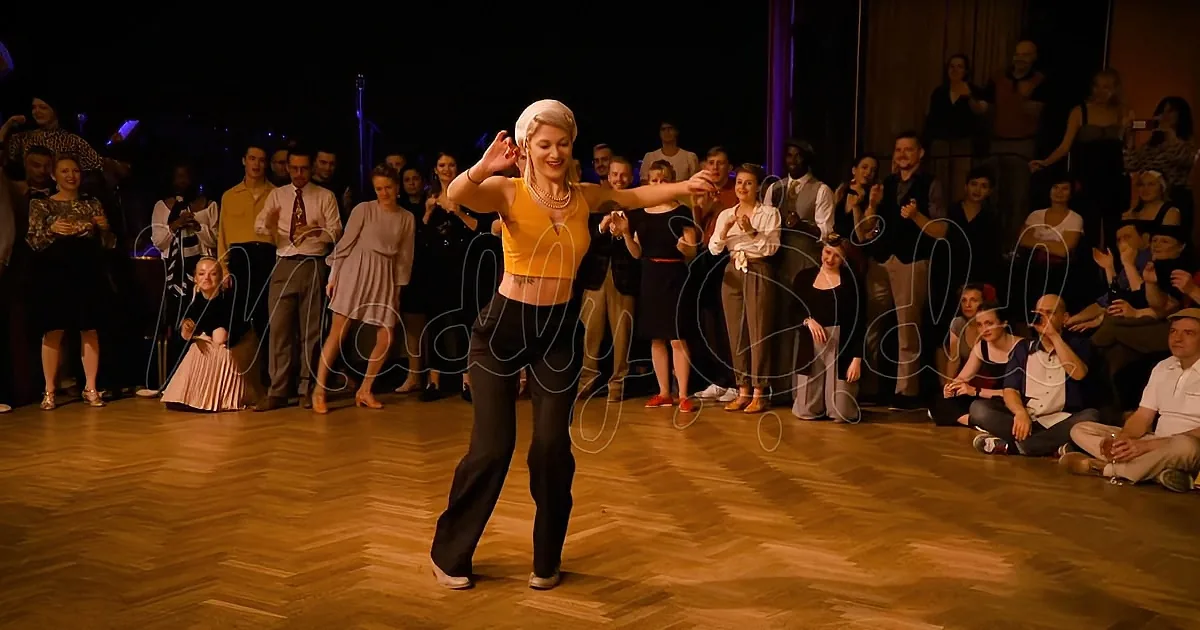 A woman in a yellow top and black pants dances on a wooden floor, surrounded by an audience in a dimly lit room.