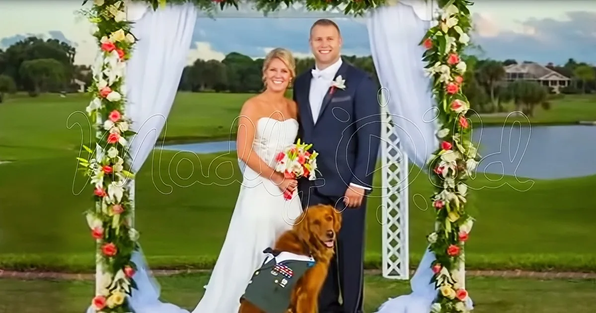 A bride and groom stand under a floral arch with a dog in a military uniform.