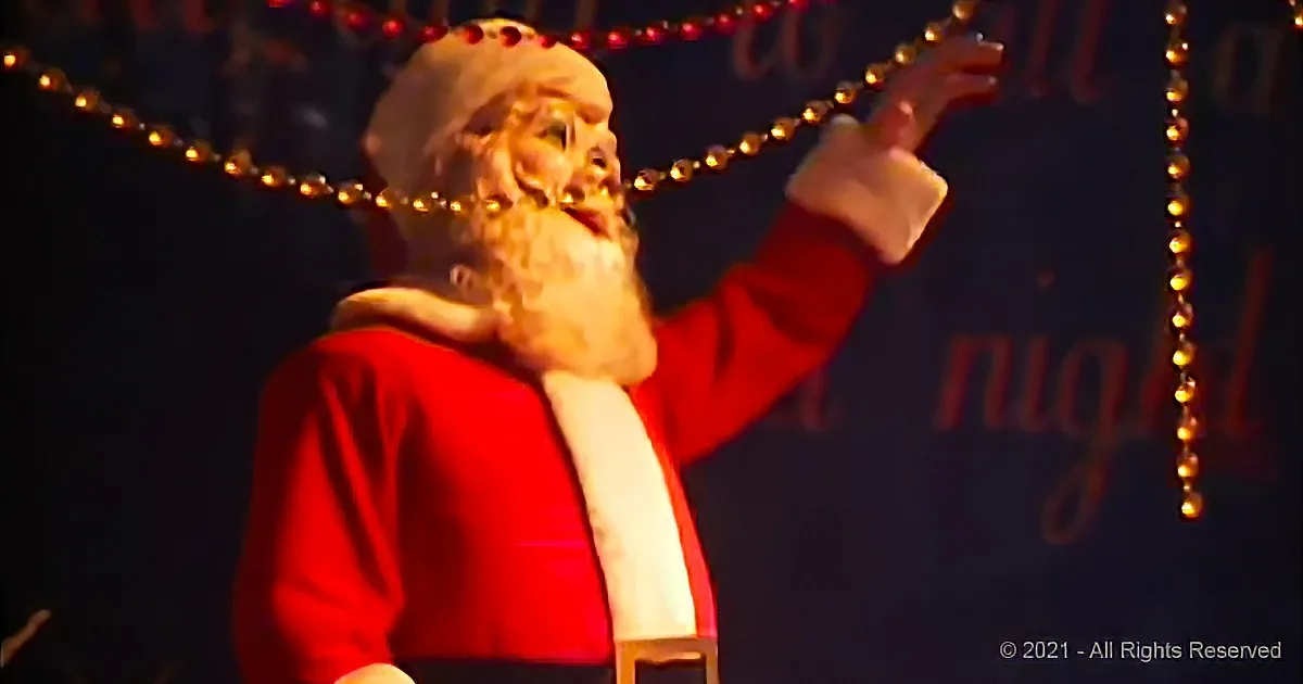 A person dressed as Santa Claus with a red suit and white beard, raising one arm.