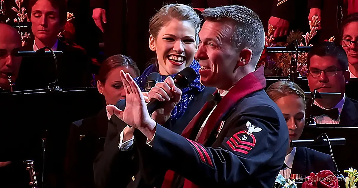 A man in a military uniform holds a microphone, smiling, with a woman beside him, also smiling, in a formal setting.